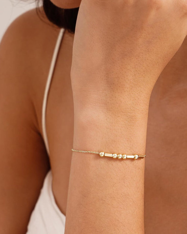 A delicate gold morse code chain bracelet with three small polished bead-like accents worn on a woman's wrist, shown in a close-up lifestyle shot against a soft beige background.