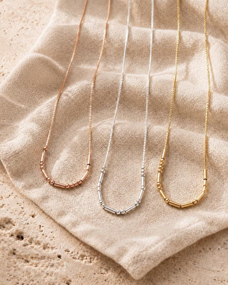 Blancora Gold, Silver and Rose Gold -  delicate necklace pendants in rose gold, silver, and yellow gold, laid out on a textured beige fabric surface.